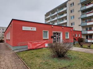 Ein rotes gebäude steht im Vordergrund, an einem Geländer befindet sich ein Plakat mit der Aufschrift Corona-Teststation, im Hintergrund ein großer Wohnblock in grau mit vielen Etagen.