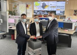 Drei Männer in einem Raum voller Computerarbeitsplätze drücken auf einen großen Knopf