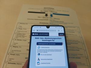 Ein Wahlschein liegt auf einem Tisch, darüber ein Smartfone mit der geöffneten Seite um die Briefwahl zu beantragen.