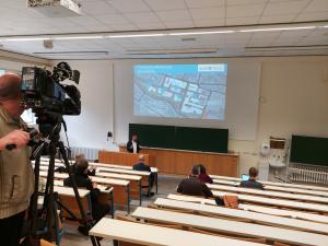 Eine Person steht vor einer Präsentation, mehrere Personen sitzen im Publikum. Auch ein Redakteur mit einer Kamera ist anwesend.