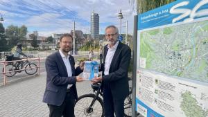 Zwei Männer halten eine Urkunde in die Kamera, im Hintergrund Jena