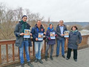 5 Menschen halten Plakate in die Kamera, hinter ihnen ein Fluss