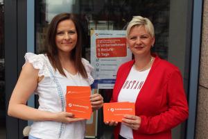 Zwei Frau stehen vor einer Glastür und schauen in die Kamera. Sie halten Flyer in der Hand. 