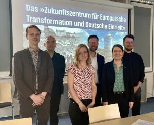 Vier Männer und zwei Frauen stehen versetzt nebeneinander vor einer Wand und schauen in die Kamera