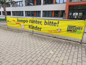 Vor einer Schule hängt ein Banner, dass zu Vorsicht im Verkehr aufruft.