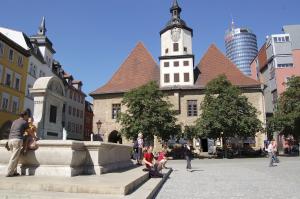 Im Vordergrund ein steinerner Brunnen auf einem großen Platz, im Hintergund mehrere Gebäude.