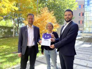 Drei Menschen halten eine Urkunde in die Kamera