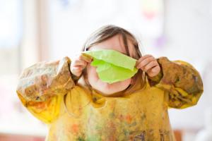 EIn kleines Mädchen hält sich ein Stück Papier vor das Gesicht.