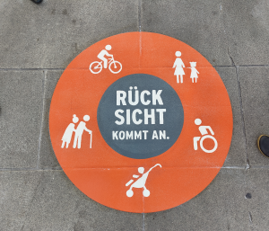 Bordenmarkierung: Orangerkreis mit weißen Symbolen (Rollstuhlfahrer, Senioren, Mutter mit Kind, KInderwagen, Radfahrer) in der Mitte steht "Rücksicht kommt an"