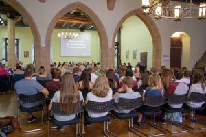 Viele Menschen sitzen in einem Saal auf Stühlen und sehen sich eine Präsentation an.
