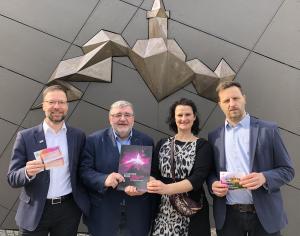 Drei Männer und eine Frau stehen vor einer Wand der Leuchtenburg und halten Karten für die "Tage der Jenaer" in der Hand
