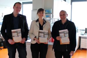 Drei Menschen halten eine Broschüre in die Kamera