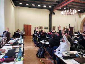 Mitglieder des Stadtrates sitzen im Historischen Rathaus an Tischen und stimmen über einen Beschluss ab. Viele Mitgliedder heben die Hände.