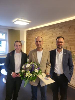 3 Personen stehen in einem Raum, eine Person hält Blumen und eine Urkunde in der Hand.