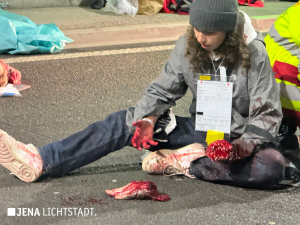 Eine Statistin sitzt auf dem Boden. Ihr einer Arm ist Blut verschmiert und als Armstumpf dargestellt. Vor ihr liegt eine realistisch aussehende künstliche Hand.