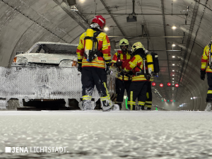 Feuerwehrleute stehen neben dem mit Löschschaum bedeckten Auto und löschen im Auotinneren die letzten Flammen.
