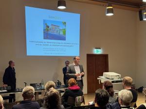 Bürgermeister Christian Gerlitz stehtim Plenarsaal im Historischen Rathaus und spreicht vor sitzenden Personen. HInter ihm ist eine Präsentation via Beamer an die Wand geworfen zum Thema Rahmenplanung zur Weiterentwicklung des Bahnhofes Jena West