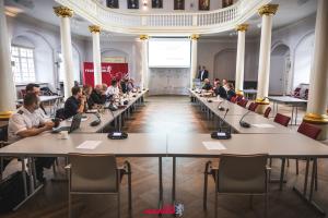 Bild zeigt großen Tisch in U-Form in einem großen Saal, an dem viele Menschen sitzen.