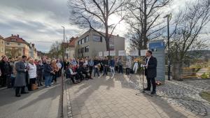 An einer Gedenkstele auf dem Gehweg vor dem Angergymnasium steht OB Thomas NItzsche und hält an einem Mikrofon eine Rede. Vor der Stele liegen Blumen, damit steht ein Kranz auf einem Ständer. Im Halbkreis um die Stele stehen Menschen unterschiedlichen Alters und höheren zu, einige sitzen auf Stühlen.