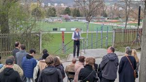 Ein junge Person liest stehend an einem Mikrofon etwas vor. Vor ihm stehen Mensche im Halbkreis und hören zu. Hinter ihm ist ein Sportplatz zu sehen.