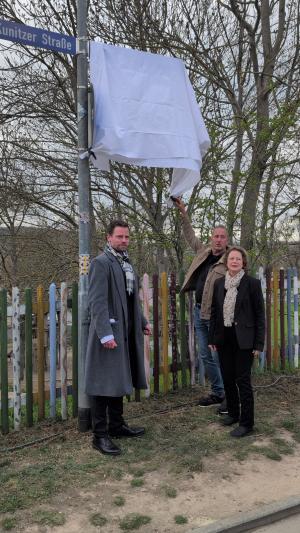 Zwei Männer und eine Frau stehen unter einem mit einem weißen Tuch verhüllten Straßenschild. Der eine Mann hat das Tuch an einer Ecke in der Hand.