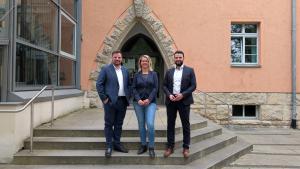 Zwei Männer und eine Frau stehen nebeneinander auf der Treppe, die zum Haupteingangs des technihschen Rathauses in Jena führt.