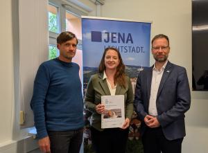 Zwei Männer und eine Frau stehen nebeneinander vor einem Roll-Up der Stadt Jena.