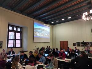 Menschen sitzen an Tischen in einem historischen Gebäude, im Vordergrund steht ein Mann an einem Rednerpult