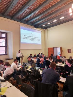 Menschen sitzen an Tischen in einem historischen Gebäude, im Vordergrund steht ein Mann an einem Rednerpult