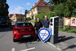Eine Frau und ein Mann stehen zwischen einem Auto und einer E-Ladesäule. Die Frau hält ein Ladekabel in der Hand und der Mann ein Schild mit der Aufschrift: Carsharing Standort teilAuto.