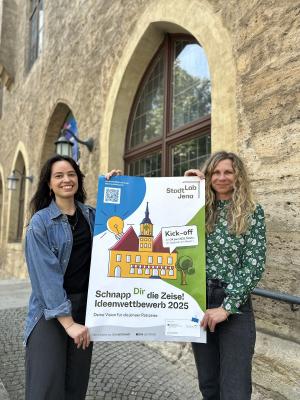 2 Frauen halten vor StadtLab Veranstaltungsplakat hoch