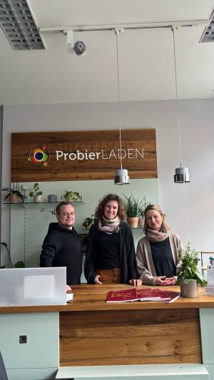 Ein Mann und zwei Frauen stehen hinter einer Empfangstheke. Dahinter hängt ein Bild mit der Aufschrift ProbierLaden.