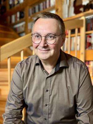 Ein Mann um die 50 sitzt vor einer Holztreppe, trägt eine Brille und ein hellbraunes Hemd.