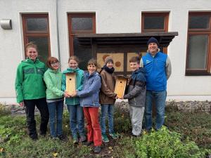 Fünf Jugendliche (drei Mädchen und zwei Jungen) halten zwei Nistkästen aus Holz. Links neben ihnen steht eine Frau und auf der rechte Seite ein Mann.