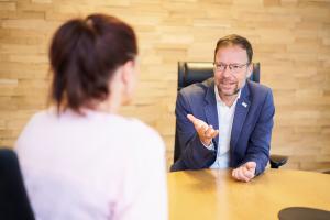 Ein Mann und eine Frau sitzen an einem Tisch und unterhalten sich. Sie ist nur von hinten zu sehen.