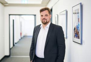 Mann in blauem Anzug steht in einem Flur mit Bildern an der Wand und lächelt