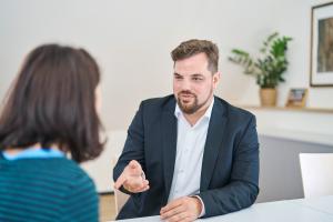 Mann redet mit einer Frau an einem Tisch, sie ist von hinten zu sehen