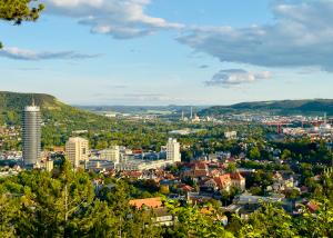 Das Foto zeigt eine weite Panoramaaufnahme der Stadt Jena in Thüringen, aufgenommen vom Landgrafenblick aus. Im Vordergrund sind grüne Baumkronen zu sehen, dahinter breitet sich die Stadt mit ihren Wohnhäusern und Bürogebäuden aus. Links ragt der runde Jentower deutlich über die übrigen Gebäude hinaus. Im Hintergrund ziehen sich die bewaldeten Hügel des Saaletals entlang, und der Himmel ist leicht bewölkt mit Sonnenlicht, das die Landschaft in warmes, freundliches Licht taucht.