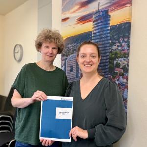 Zwei Frauen schauen freundlich in die Kamera und halten eine Broschüre