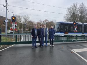 Vier Männer stehen auf der Straße vor einer geschlossenen Schranke. Im Hintergrund fährt eine Straßenbahn vorbei. 