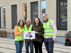 Vier Personen stehen nebeneinander vor einem modernen Gebäude mit großen Fenstern. Drei von ihnen tragen leuchtend gelbe Warnwesten, eine Person in der Mitte trägt eine dunkelgrüne Jacke. Die beiden Personen links halten gemeinsam ein Foto einer Rikscha, die für das Projekt »Radeln ohne Alter« vorgesehen ist. Alle lächeln in die Kamera.
