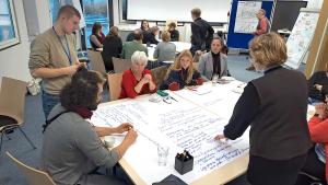 In einem hellen Seminarraum sitzen mehrere Gruppen von Menschen an Tischen und arbeiten gemeinsam an großen Papierbögen. Im Vordergrund beugen sich Teilnehmende über ein großflächiges Blatt, auf dem viele handgeschriebene Stichpunkte stehen. Eine Frau zeigt auf einzelne Notizen, während andere aufmerksam zuhören. Im Hintergrund sind weitere Kleingruppen im Gespräch, Moderationsmaterialien und beschriebene Flipcharts sichtbar. Die Stimmung wirkt konzentriert und kooperativ.