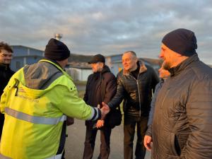 Eine Person in einer gelben Jacke der Stadtwerke Jena gibt mehreren Besuchern im Freien die Hand. Die Gruppe steht lächelnd beisammen, im Hintergrund sind Gebäude und ein bewölkter Himmel zu sehen, das Licht der tiefstehenden Sonne fällt warm auf die Szene.