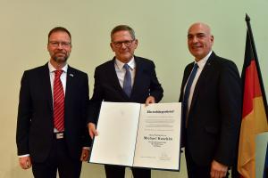 Drei Männer stehen nebeneinander, einer hält ein großes Buch in de Hand.