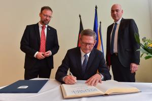 Ein Mann unterschreibt in einem Buch, zwei Männer stehen hinter ihm und schauen dabei zu.