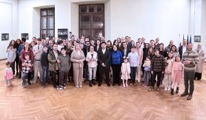 Große Gruppe von Menschen unterschiedlichen Alters steht gemeinsam in einem hellen Saal mit Parkettboden und hohen Fenstern für ein Gruppenfoto.