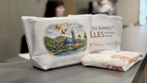 Zwei weiße Stofftaschen mit buntem Aufdruck stehen auf einem Tisch, daneben liegen Windeln; im Hintergrund unscharf Personen in einem Innenraum.