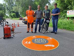 Drei Männer und eine Frau mit Fahrrad stehen hinter einer runden Markierung auf der Straße.
