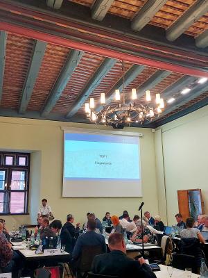 Im historischen Rathaus steht eine Person an einem Rednerpult und andere Menschen sitzen an Tischen, im Hintergrund ist eine Präsentation an die Wand projiziert, darauf steht das Wort "Fragestunde"