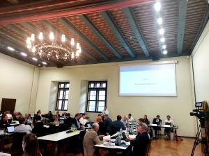 Menschen sitzen an Tischen im Historischen Rathaus, ein Mann steht an einem Rednerpult, im Hintergrund ist eine Präsentation an die Wand geworfen.